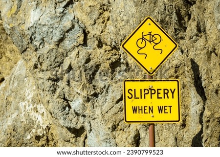 Two yellow SLIPPERY WHEN WET warning signs near a rocky cliff face. Yellow caution sign warns cyclists of slippery road new a rocky outcropping