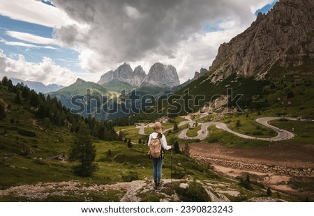 The back of an Asian female hiker stood atop a rock in front of an awesome view of a mountain pass snaking up the lush green valley with rugged mountain ranges in view in the diatance