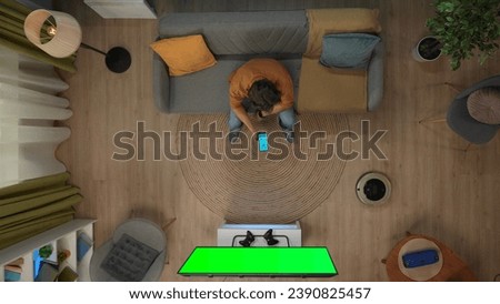 Top view of man sitting on the sofa holding smartphone looking at app for gadgets, tv with chroma key green screen. Royalty-Free Stock Photo #2390825457