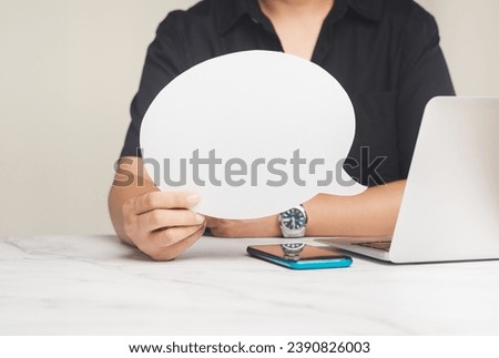 A young man in a black short-sleeved shirt holding a blank white speech bubble while working at a desk. Close-up photo. Speech bubble concept.