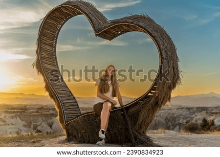 A young woman in the mountains at sunset sits on a bench shaped like a heart made of branches, with a canyon in the background.