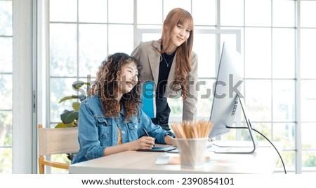 Asian professional successful young female creative graphic designer in casual outfit sitting smiling drawing sketching artwork with stylus pen when colleague helping advising pointing pc monitor.