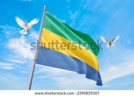 Waving flag of Gabon in beautiful sky and flying pigeons. Gabon flag for independence day. The symbol of the state on wavy fabric. Royalty-Free Stock Photo #2390859507