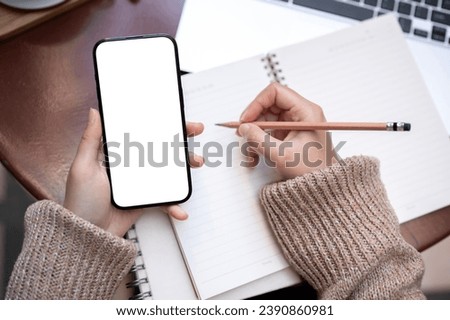 Close-up image of a woman is taking notes in her notebook and using her smartphone while sitting at a table outdoors. A white-screen smartphone mockup in a woman's hand.