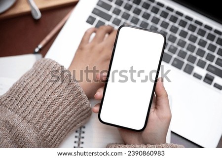 Top view image of a woman working on her laptop computer and using her smartphone at a table in a cafe. A white-screen smartphone mockup in a woman's hand.