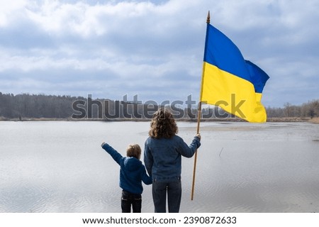 boy and young woman, younger brother and adult sister, stand next to each other holding hands with large flag of Ukraine. Back view. Family, unity, refugees, support. day of dignity and freedom Royalty-Free Stock Photo #2390872633