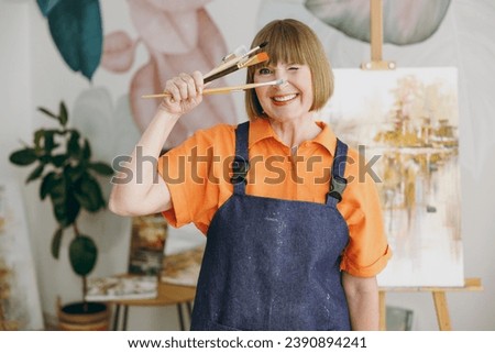 Elderly cheerful artist woman 50 year old wears casual clothes stand near easel with painting artwork hold cover eyes through brushes spend free spare time in living room indoor. Leisure hobby concept