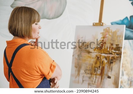 Elderly depressed minded pensive artist woman 50 years old wears casual clothes stand near easel with painting artwork prop up chin spend free spare time in living room indoor. Leisure hobby concept