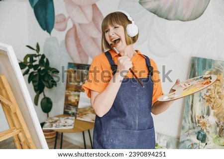 Elderly artist woman 50 years old wear casual clothes stand near easel with artwork paint listen music in headphones sing song in microphone spend free time in living room indoor Leisure hobby concept Royalty-Free Stock Photo #2390894251