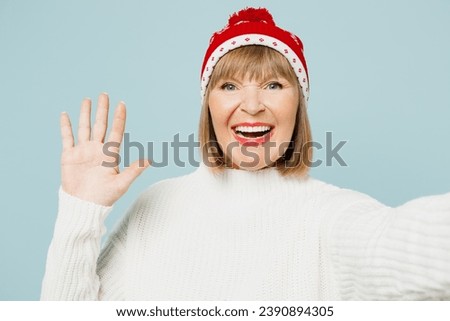 Close up merry elderly woman 50s years old wear sweater red hat posing doing selfie shot on mobile cell phone waving hand isolated on plain blue background. Happy New Year celebration holiday concept