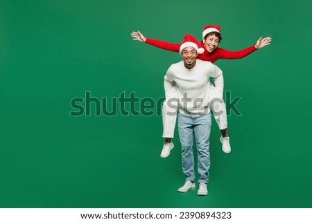 Full body merry young couple man woman wear red casual clothes Santa hat posing giving piggyback ride to joyful, sit on back isolated on plain green background. Happy New Year 2024 Christmas concept