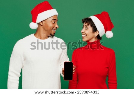 Merry smiling young couple man woman wear red casual clothes Santa hat posing hold use blank screen mobile cell phone isolated on plain green background. Happy New Year 2024 Christmas holiday concept