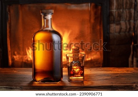 Glass of whiskey and whiskey bottle on old wooden table and blurred fireplace at the background.