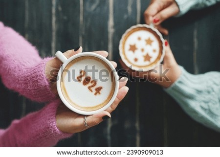 Coffee cup with the number 2024 on frothy surface in female hands holding over blurred dark wooden table and another one with star symbols on frothy surface. Happy new year 2024 food art theme. Royalty-Free Stock Photo #2390909013