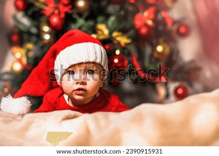 Joyful baby looking at camera in Christmas photo. Newborn baby in red cap of Santa Claus and Christmas tree in background