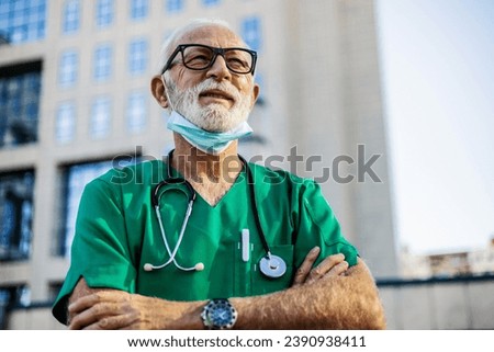 Portrait of a successful mature doctor with a beard and in a medical uniform, outside a modern clinic, a hospital director with crossed arms smiling and looking away.