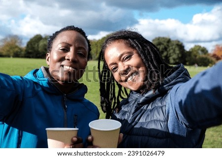 Happy sport mature women drinking coffee and making a selfie outdoors. It is a sunny autumn day and they are wearing sport winter outfits. They have short hair and dreadlocks. Royalty-Free Stock Photo #2391026479