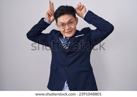 Young asian man wearing business suit and tie posing funny and crazy with fingers on head as bunny ears, smiling cheerful 