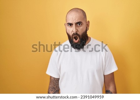 Young hispanic man with beard and tattoos standing over yellow background in shock face, looking skeptical and sarcastic, surprised with open mouth 