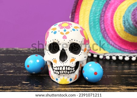 Painted human skull for Mexico's Day of the Dead (El Dia de Muertos) with maracas and sombrero on wooden table against purple background