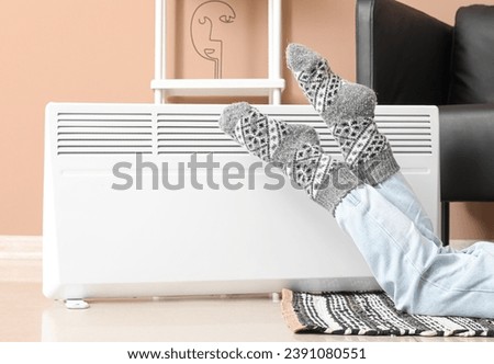 Young woman in warm socks near electric heater at home. Concept of heating season