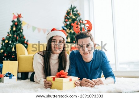 Young happy Asian woman wearing a Santa Claus hat with her boyfriend with a Christmas gift while lying down on the carpet and looking at the camera with a Christmas tree in the background.