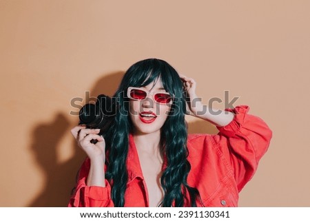 Cheeky shouting bright fashion girl with dark green long hair, bright red lips, top and sunglasses holding retro film camera on beige background. Trendy young hipster photographer, travel photo.
