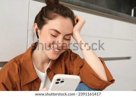 Portrait of happy young beautiful woman, listening music in wireless earphones, using smartphone, watching videos on mobile phone with headphones on. Royalty-Free Stock Photo #2391161417