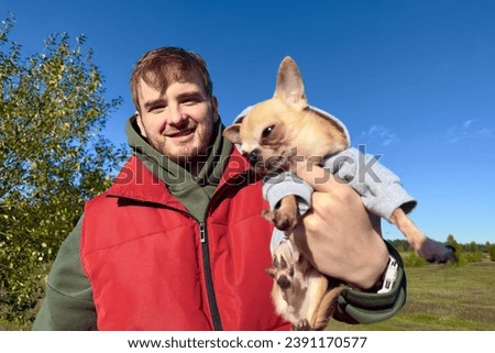 Portrait of happy young man with his Chihuahua dog, hold on hands little puppy on natural background in sunny forest or park.