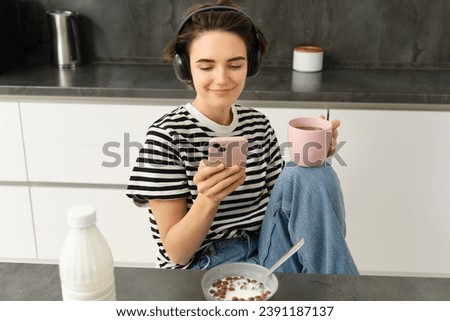 Portrait of young woman drinking tea and listening to music in headphones, scrolling social media while having lunch in kitchen, smiling happily. Royalty-Free Stock Photo #2391187137