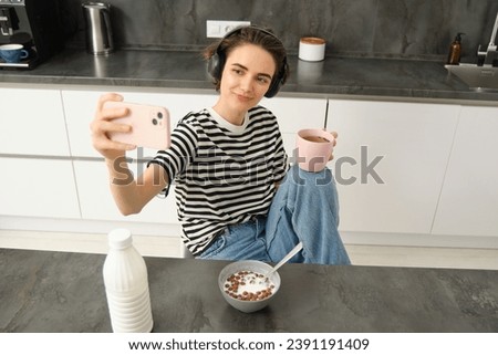 Stylish young woman, student taking selfie in the kitchen, drinking tea and eating breakfast, making pictures for social media app.