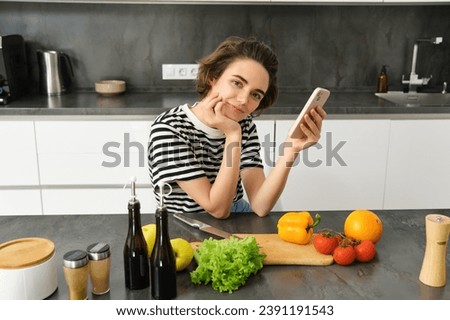 Portrait of cute brunette woman in the kitchen, cooking, searching recipe on smartphone, taking pictures for her food blog, cooking meal from vegetables, vegan salad.