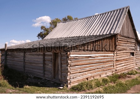 Historic Mormon Row houses, barns and outbuildings surrounded by green and brown scrub brush and grass.  Background against the Grand Teton mountains and blue cloud skies.  Historical West settlement.