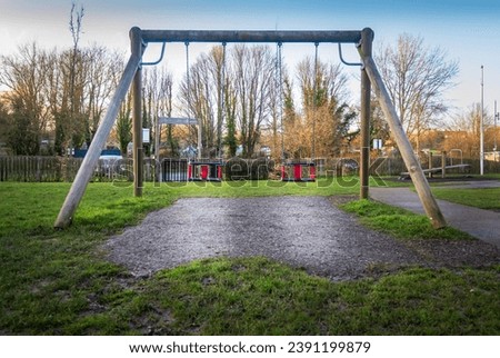 Swingset in outdoor children's playground park. Two swings, metal chain, wooden support beams. Fun childhood activity equipment for kid's in public play area. Blue sky, daytime, late afternoon. Royalty-Free Stock Photo #2391199879