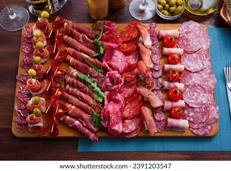 Top view of traditional Spanish meat platter - sliced dry-cured jamon, bacon and sausages on wooden board with olives, tomatoes and greens Royalty-Free Stock Photo #2391203547