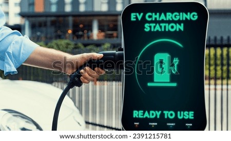 Businessman pull and hold EV charger plug form electric car charging station at city car park area background. Futuristic clean sustainable energy and EV car technological advancement.Peruse