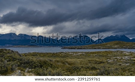 The Andes mountain range. Snow caps of peaks against the sky and clouds. Houses in the city of Ushuaia at the foot, on the banks of the Beagle Canal. An island with sparse vegetation in the foreground