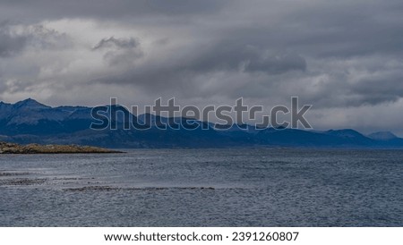A picturesque mountain range of the Andes against a background of cloudy sky and ocean. An island with sparse vegetation is visible. Argentina. Beagle Channel. Tierra del Fuego Archipelago.