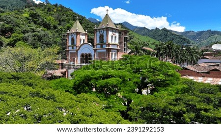 Drone view of a traditional church with twin spires amidst green foliage against mountains under a blue sky in Colombia.