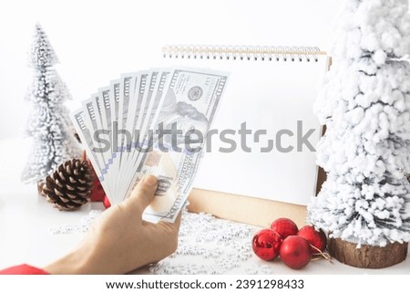 Gentle hands cradle money with a white screen mock-up and White calendar with a festive scene of a beautiful Christmas tree adorned, with Christmas balls, pine cones, and a red house gift box.
