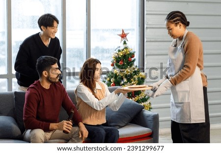 Millennial Asian Indian multinational cheerful male female friends celebrate Christmas Eve Happy New Year Chef serve hot tasty pizza pan in living room full festive decoration with Xmas tree.