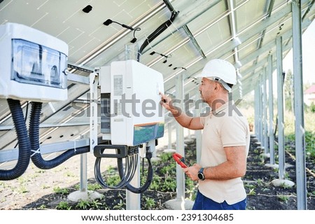 Professional worker checking voltage on solar inverter. Male using current probe to measure output voltage. Man in helmet pointing at inveter dial on back side of PV panel. Royalty-Free Stock Photo #2391304685