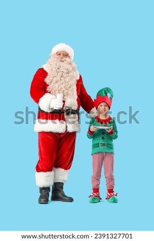 Santa Claus and cute little elf with milk and cookies on blue background