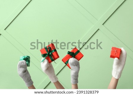 Female hands in stylish warm mittens with Christmas gifts against color wall