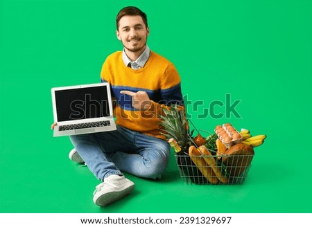 Young man with shopping basket pointing at laptop on green background