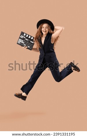 Beautiful actress with movie clapper jumping on beige background Royalty-Free Stock Photo #2391331195