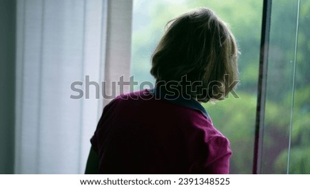 Pensive Senior woman standing by window looking out at view during rainy day. Contemplative elderly retired lady at home while raining outside, staring at rain Royalty-Free Stock Photo #2391348525