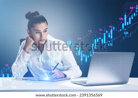 Attractive young businesswoman working at desk with laptop and glowing upward candlestick forex chart on blurry grid background. Trade, finance and money concept