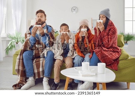 Achoo. Family of four have caught bad cold all together. Sick mom, dad and children in warm clothes and plaids sitting on couch in living room and sneezing in paper handkerchiefs. Winter, flu concept Royalty-Free Stock Photo #2391365149