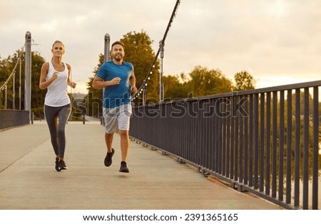 Two runners in the evening. Young boyfriend and girlfriend having a jogging workout on city streets. Workout buddies in sportswear running along the bridge fence together. Sport, exercise concept Royalty-Free Stock Photo #2391365165